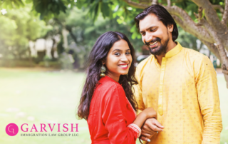 Indian Couple outside in traditional clothing. The woman is wearing red and the man is wearing yellow. They are holding hands with their arms interlocked with bodies facing each other but both looking toward the camera.