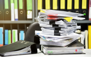 In an office, stacks of papers held together with large binder clips. Colored office binders in the background.